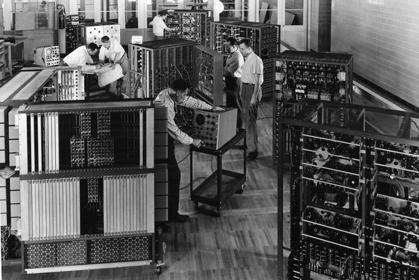 عکس تاریخی سیاهوسفید از نصب و راهاندازی سیستم کامپیوتر آیبیام ۷۰۱ (IBM 701) در دهه ۱۹۵۰، نمایش تیم مهندسان، اپراتورها و قفسههای عظیم واحدهای پردازش الکترونیکی در محیطی صنعتی.
