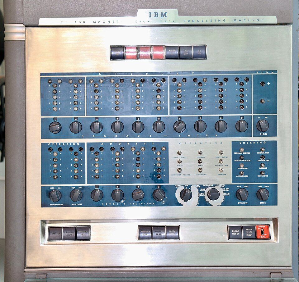 نمای نزدیک و باکیفیت از پنل کنترل اصلی رایانه قدیمی آیبیام ۶۵۰ (IBM 650) با قاب فلزی نقرهای و المانهای آبیرنگ، شامل آرایش منظم چراغهای نشانگر برای نمایش ثباتهای دیجیتال، دو ردیف کلید چرخشی برای وارد کردن دادهها و آدرسدهی، و مجموعه دکمههای فشاری راهاندازی، ریست، و مدیریت خطا در پایین پنل، که برای مدیریت ماشین پردازش داده با درام مغناطیسی طراحی شده است.